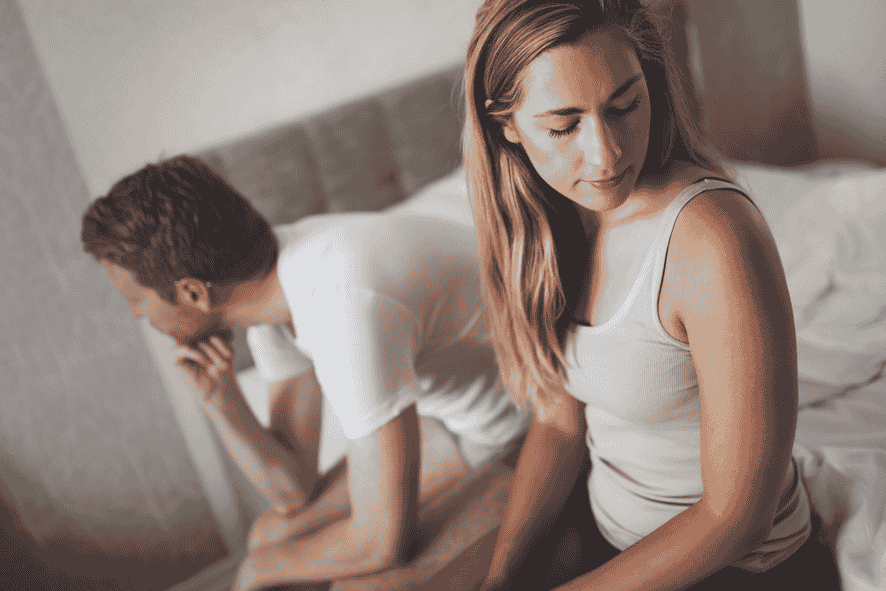 Young woman and man sitting on bed facing away from each other and looking distressed.