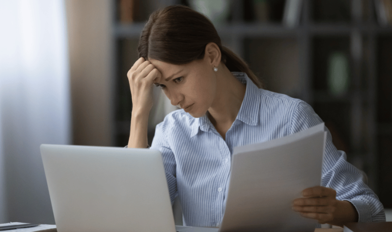middle-aged woman looking stressed sitting in front of a laptop and holding a packet of paper.