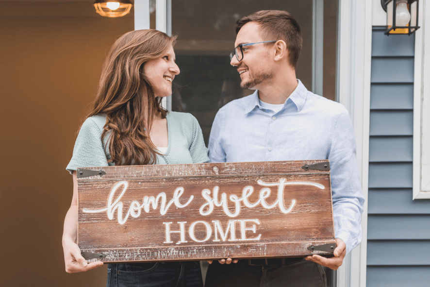 Girlfriend and boyfriend smiling at each other in front of home holding a sign together that reads "home sweet home"