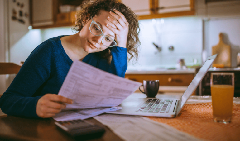 woman holding head in hand and looking stressed while looking at bill at kitchen table.
